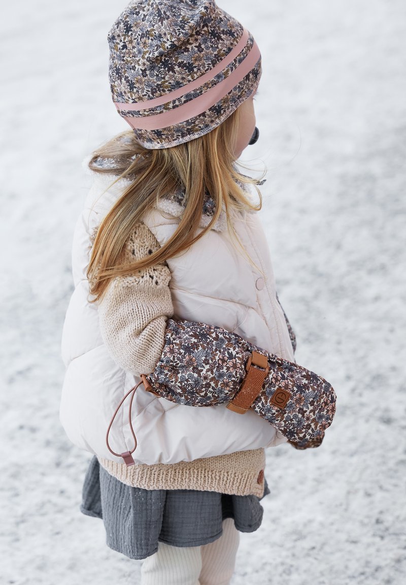 Ungt barn i vinterkläder med blommig mössa och vantar som står på snöig mark, håller vantarna bakom ryggen, vänd mot höger.