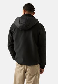 Czarna kurtka zapinana na zamek błyskawiczny z ocieplanym kapturem, regulowanymi sznureczkami i elastycznymi mankietami, w połączeniu z beżowymi spodniami. Gładka faktura materiału.