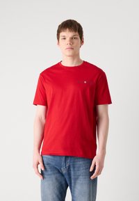 T-shirt en coton rouge à manches courtes, col rond et impression graphique sur le côté gauche de la poitrine. Associé à un jean en denim bleu.