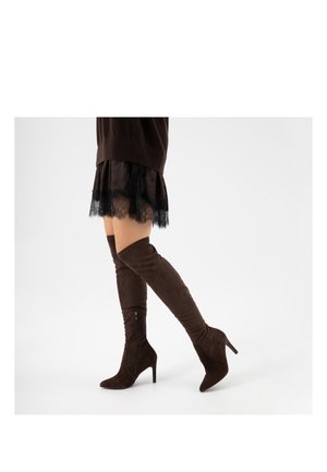 Donna che indossa una gonna di pizzo nera e stivali cuissard in camoscio marrone scuro con tacco a spillo, che cammina su sfondo bianco.