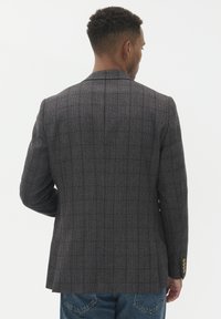 Muž otočený zády, oblečený v tmavě šedém kostkovaném saku a modrých džínách, stojící na jednoduchém bílém pozadí.