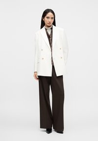 Fehér kétsoros blézer arany gombokkal, barna mintás nyakkendővel és sötét, bőszárú nadrággal párosítva; elegáns, modern dizájn.