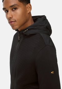 Veste à capuche noire avec des motifs hexagonaux texturés sur le devant et des manches lisses ; comprend un détail de poignet avec logo et une fermeture éclair.