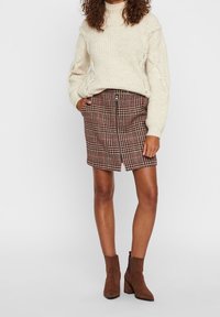 Vrouw in een crèmekleurige kabelstriktrui, een bruin geruite rok met rits en bruin suède enkellaarsjes, staand met een hand in haar zak.