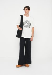 Weißes Baumwoll-T-Shirt mit schwarzem Grafikdesign, lockere Passform; kombiniert mit schwarzen weiten Hosen und hellbraunen Clogs, tragt eine schwarze Umhängetasche.