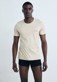 T-shirt en coton beige avec un col rond et des manches courtes, arborant un logo rouge "HUGO". Le modèle porte un short noir, se tenant devant un fond clair.