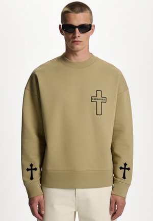 Jeune homme portant des lunettes de soleil et un sweat-shirt beige avec des croix noires, une sur la poitrine avec le texte "Karma Kills" et une sur chaque manche.