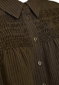 Brūns pogu krekls ar vertikālām svītrām, ar smocka detaļām uz ķermeņa un apaļām brūnām pogām. Gluda tekstūra ar apkakli.