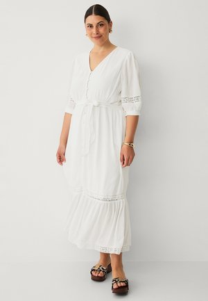 Vrouw die een witte midi-jurk met knopen en kanten details draagt, driekwart mouwen, een taillekoord, gouden sieraden en sandalen met luipaardprint.