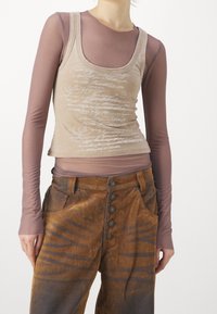 Beige linnenmask med vit skript-design lagd över en genomskinlig, ljusbrun långärmad skjorta och bruna mönstrade höga byxor med knappar.