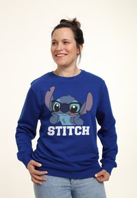Blaues Sweatshirt mit einem Cartoon-Charakter mit großen Ohren und Brille, Text "STITCH" in Weiß, aus weichem Stoff, mit langen Ärmeln.