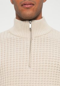 Beige strikket genser med høy krage og halv glidelås foran. Teksturert mønster gjennomgående med en glatt glidelås.