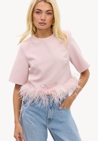 Femme blonde portant un haut rose pâle à manches courtes avec un bord en plumes à l'ourlet, associé à un jean taille haute bleu clair, main dans la poche.