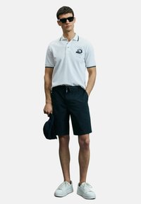 Man in een witte poloshirt, marineblauwe shorts, witte sneakers, zwarte zonnebril, een marineblauw pet in de hand, staand tegen een effen achtergrond.