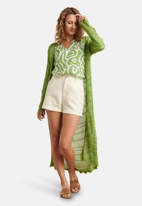 Langer, offen gestrickter grüner Cardigan, gemustertes grün-weißes Oberteil, cremefarbene Shorts und braune Slide-Sandalen. Leichtes, lässiges Outfit.