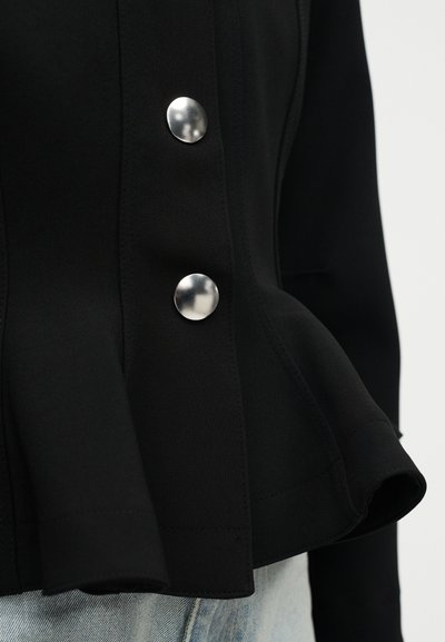 Μαύρο εφαρμοστό σακάκι με βολάν στη μέση και δύο μεγάλα λαμπερά ασημένια στρογγυλά κουμπιά, φορεμένο πάνω από ανοιχτόχρωμο τζιν παντελόνι.