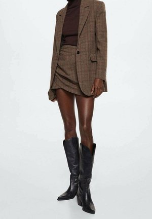 Personne portant un blazer à carreaux marron et une jupe assortie, un haut marron foncé et des bottes en cuir noires à talons mi-mollet, debout sur fond blanc.