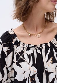 Złota naszyjnik z wieloma zawieszkami, w tym sercem i splątanymi kształtami, noszony na czarno-białej bluzce w kwiatowy wzór.