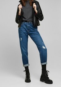 Blauwe spijkerbroek met een relaxte pasvorm, voorzien van opgerolde manchetten en versleten patches. Gepaard met een grijze shirt en een zwarte leren jack. Zwarte laarzen maken de look compleet.