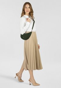 Bolso bandolera de ante verde con forma curva; modelo con blusa blanca y falda midi plisada beige, combinado con tacones nude.