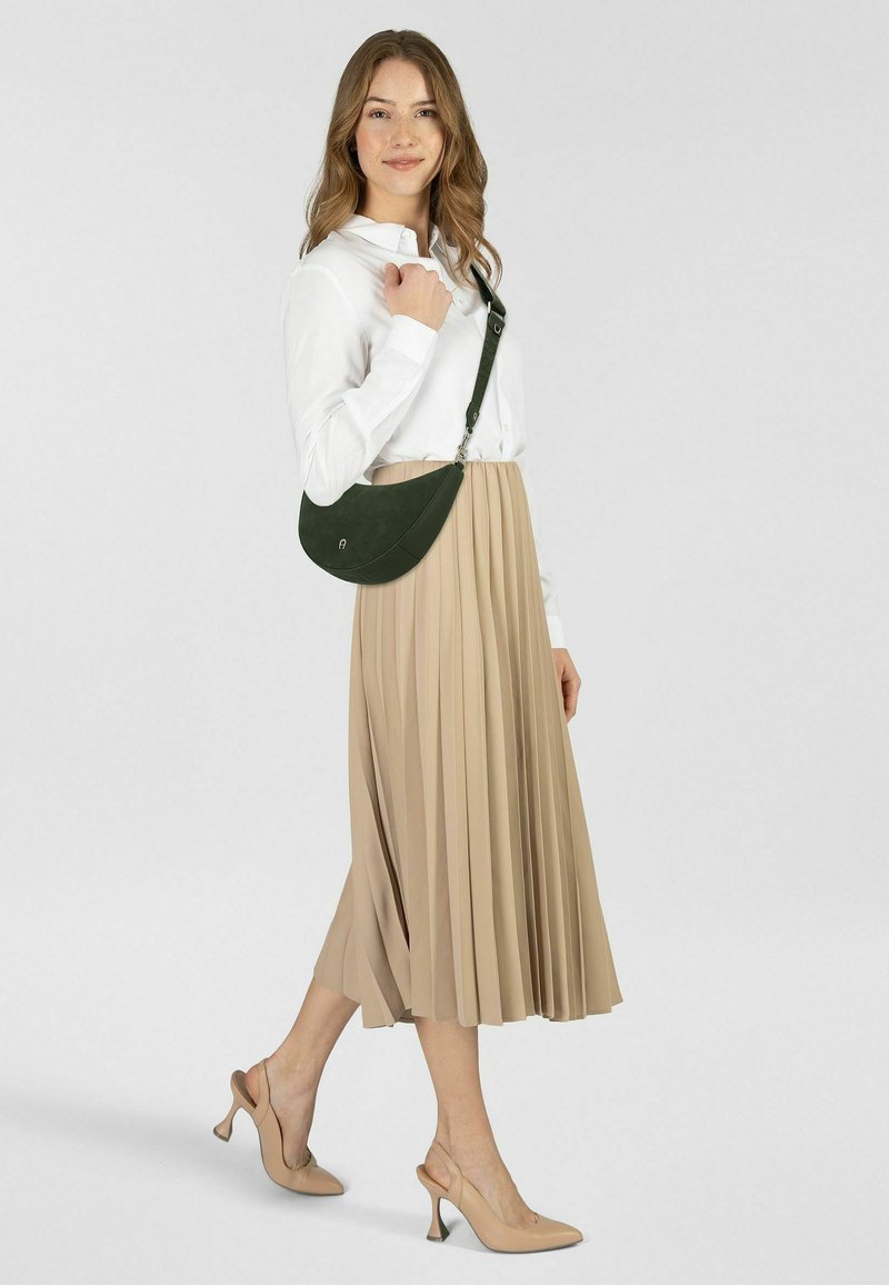 Bolso bandolera de ante verde con forma curva; modelo con blusa blanca y falda midi plisada beige, combinado con tacones nude.