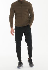 Man wearing an olive green quilted jacket, black jogger pants, and gray running shoes walking with one hand in pocket.