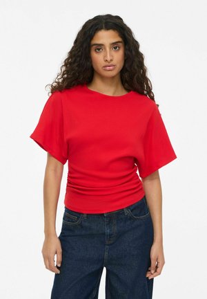 Femme aux longs cheveux bouclés portant un haut rouge vif à manches courtes et un jean bleu foncé, debout devant un fond blanc uni.