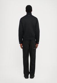 Jachetă neagră cu guler înalt și tiv elastic, asociată cu pantaloni cargo negri dotati cu buzunare laterale și fermoare. Design simplu și elegant.