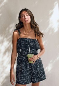 Femeie îmbrăcată într-un romper alb-negru cu buline, ținând un smoothie verde cu pai, stând în fața unui perete deschis la culoare cu modele de umbre.