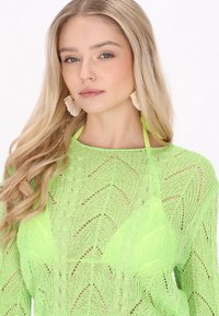 Ljusgrön, stickad tröja med ett chevronmönster och öppen väv. Har en vid necklinje och är lagd över en gul bikinöverdel.