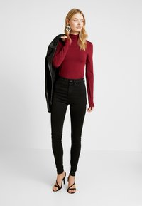 Bordeaux lange mouwen coltrui, zwarte high-waisted skinny jeans, zwarte gestileerde sandaalhakken, houdend een zwarte leren jas. Gladde stoftextuur.