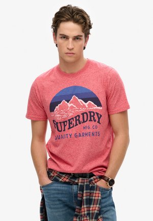 Rød meleret T-shirt med et blåt og hvide bjerggrafik, med "SUPERDRY" tekst. Båret med en ternet skjorte bundet om taljen.