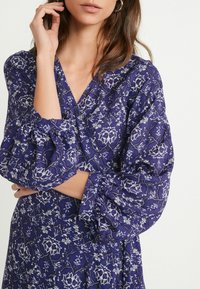 Fioletowa sukienka w kwiatowy wzór, z bufiastymi rękawami i dekoltem w kształcie litery V. Materiał wydaje się lekki z teksturowym wykończeniem.