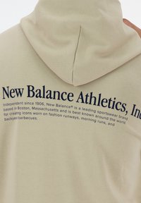 Béžová mikina s kapucí s tmavě modrým nápisem „New Balance Athletics, Inc.“ Měkká, strukturovaná látka; kapuce má šev uprostřed.