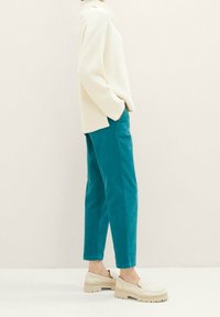 Personne portant un pull côtelé crème, un pantalon teal longueur cheville, et des mocassins épais crème, se tenant avec les mains dans les poches.