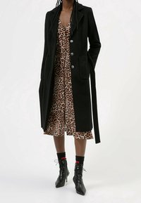Manteau long noir au design ajusté, avec des boutons argentés et des poches. Porté sur une robe à motifs léopard, associé à des bottines noires.