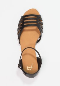 Black strappy sandal with a flat sole, featuring a tan footbed and an ankle strap with a silver buckle, designed with open sides.