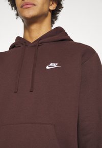 Brun hoodie i mjukt tyg med en känguruficka och en vit Nike-logga på bröstet. Dragsko på huvan.