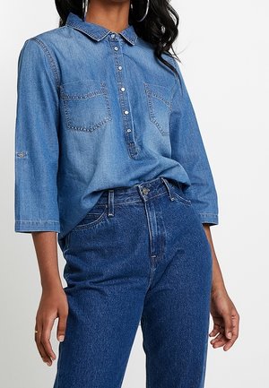 Femme portant une chemise en jean bleu moyen à boutons avec manches trois-quarts et un jean taille haute bleu foncé, debout devant un fond uni.