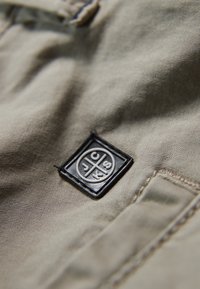 Beige kangasta, jossa on hienovaraista tekstiä, ja musta suorakulmainen etiketti, johon on kohokuvioitu kirjaimet "JCKS" sekä pyöreä muotoilu.