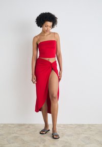 Top corto rojo ajustado con tirantes finos, combinado con una falda larga anudada que presenta una alta apertura, llevada con sandalias oscuras tipo slide. Textura de tela suave.