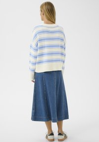 Femme aux longs cheveux blonds portant un pull rayé bleu et blanc, une jupe en jean mi-longue et des baskets blanches, debout de dos.