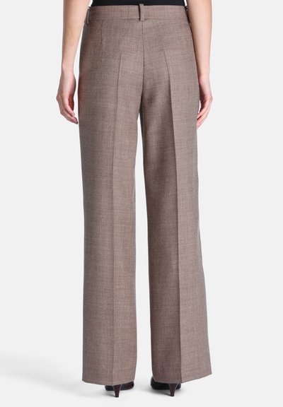 Personne portant un pantalon marron taille haute et jambes larges, debout les mains détendues le long du corps, vue de dos sur fond blanc.