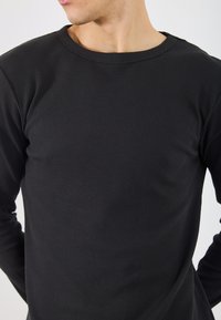 Camisa preta de manga longa feita de tecido canelado, com decote redondo e silhueta ajustada. A textura é proeminente com um padrão visível.