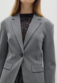 Grijze op maat gemaakte blazer met gegroefde revers, enkele knoopsluiting en twee voorzakken. Draagt over een zwarte kanten top.