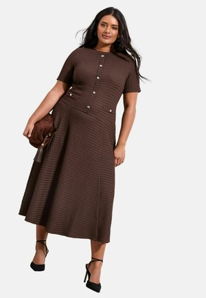 Naine, kes kannab pruuni tekstuurse lühikeste varrukatega midi-kleit nööpidega, hoides pruuni käekotti ja kandes musti pahkluu rihmadega kontsakingi.