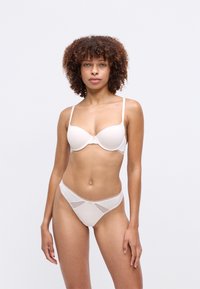 Femme aux cheveux bouclés portant un soutien-gorge beige clair et des sous-vêtements assortis, se tenant devant un fond blanc uni.