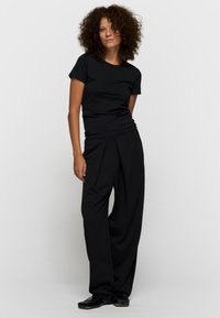 Mujer con cabello rizado que lleva una camiseta negra ajustada de manga corta, pantalones negros sueltos y zapatos negros brillantes, de pie contra un fondo liso.