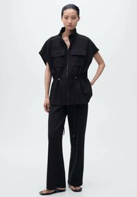 Giacca nera con colletto alto, chiusura a zip e due tasche sul petto, abbinata a pantaloni neri a gamba larga. Il materiale ha una texture liscia.