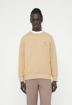 Sweat-shirt beige à col rond et poignets côtelés, avec un petit logo brodé sur la poitrine. Associé à un pantalon fuselé marron clair.